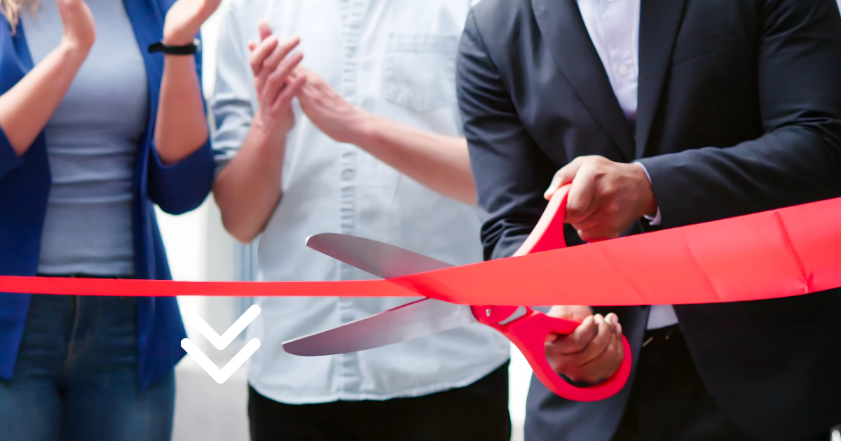 A person in a suit cuts a red ribbon with large red-handled scissors, while others clap in the background.