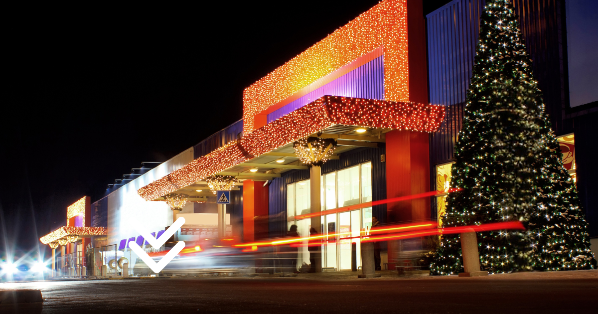A brightly lit building decorated with Christmas lights at night, with a large decorated Christmas tree on the right and light trails from passing vehicles in the foreground.
