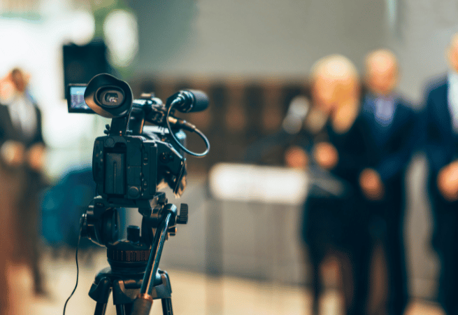 A professional video camera on a tripod is in the foreground, with a blurred group of people in suits in the background, suggesting a press conference or media event.