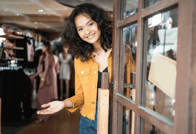 A person wearing a mustard yellow jacket and blue jeans opens a wooden-framed glass door, gesturing welcomingly into a boutique with clothing racks visible inside.